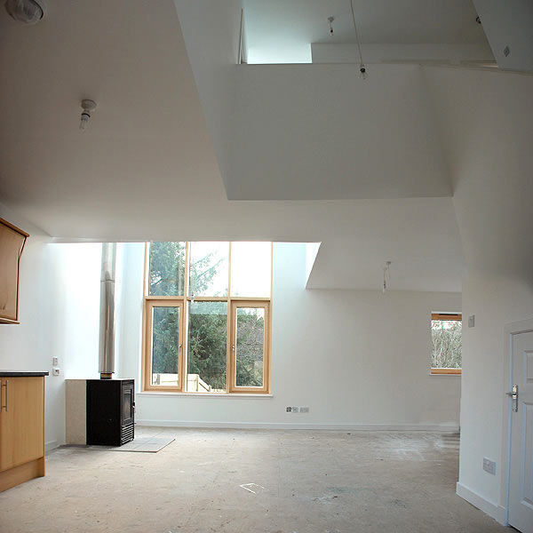 slightly distorted internal view showing the double height living area from the Kitchen and the 'bridge' across the living area at first floor leading to the third bedroom. Nearly complete but still lacking a floor finish.